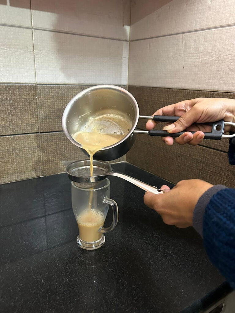 A hand holds a pot of chai while the other holds a strainer above a mug at home.
